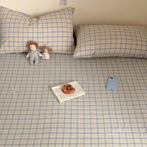 May include: A bed set featuring a light blue and beige plaid pattern on the fitted sheet and two rectangular pillows. Two small plush dolls sit on the bed, alongside a white book with a pastry on top and a light blue smartphone. Text reads Pegycrafts Copyright All rights reserved.
