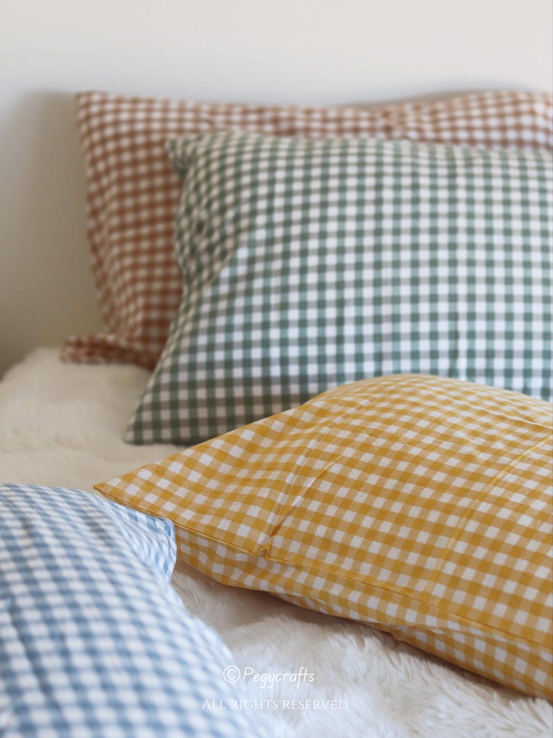 May include: Close-up of several gingham-patterned pillowcases in various colors, including yellow, green, brown, and blue. The pillows are arranged on a white, fluffy surface. The image has the text "Pegycrafts" and "ALL RIGHTS RESERVED".