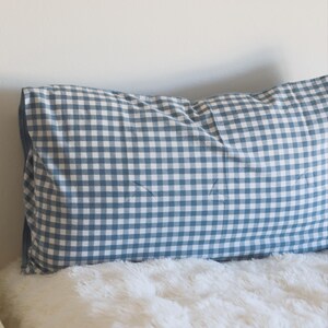 May include: A blue and white gingham patterned pillow on a white fluffy surface. The pillow is rectangular and has a classic, country-style design. The pillow is placed against a white wall.