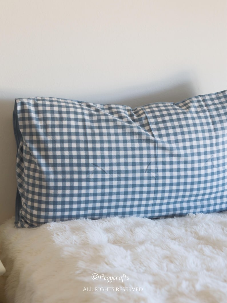 May include: A blue and white gingham patterned pillow on a white fluffy surface. The pillow is rectangular and has a classic, country-style design. The pillow is placed against a white wall.