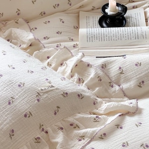 May include: A white pillowcase with a ruffled edge and a small purple flower pattern. The pillowcase has a small label that says "la vie est belle".
