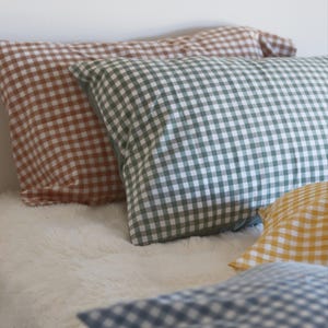 May include: Several gingham-patterned pillowcases in shades of brown, green, yellow, and blue rest on a white, fluffy surface. The pillowcases are rectangular and feature a classic checkered design. The image is well-lit, showcasing the various colors and textures.