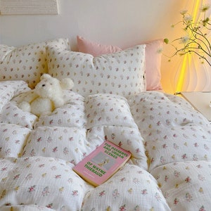 May include: A white comforter with a small floral pattern in pink, yellow, and green. The comforter is on a bed with two white pillows and a pink pillow. A book titled "The Desserts and Urban Scenes of Wayne Thiebaud" is lying on the comforter. A white teddy bear is sitting on the comforter.