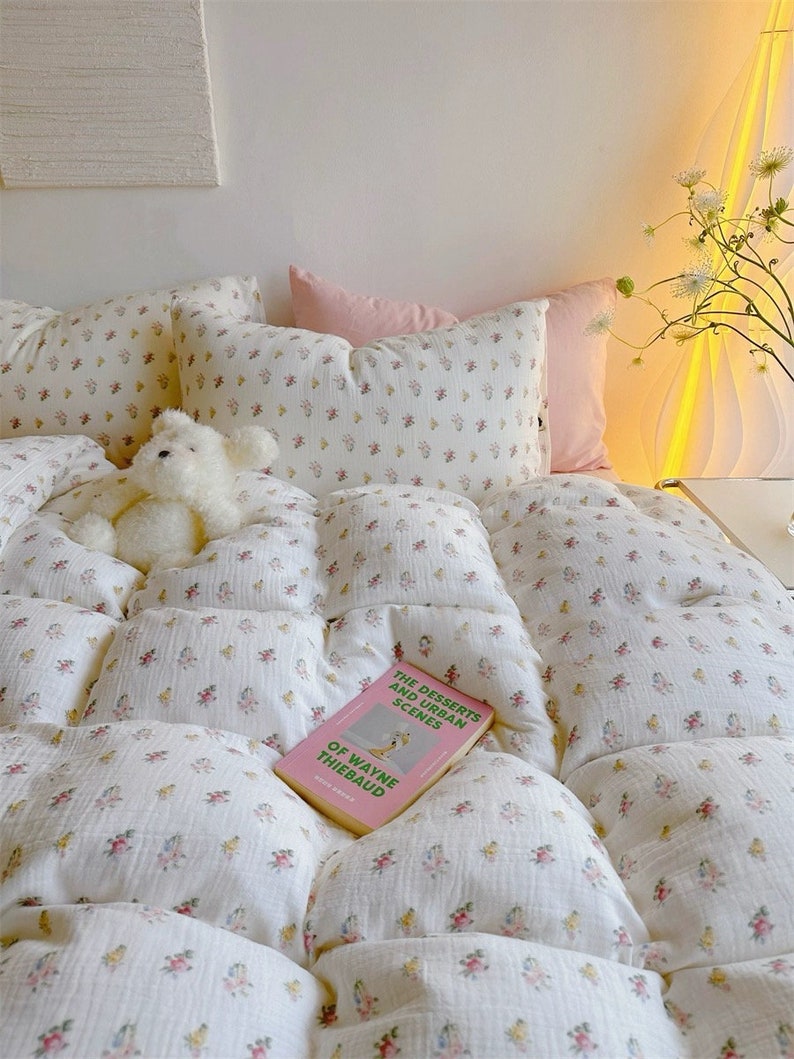 May include: A white comforter with a small floral pattern in pink, yellow, and green. The comforter is on a bed with two white pillows and a pink pillow. A book titled "The Desserts and Urban Scenes of Wayne Thiebaud" is lying on the comforter. A white teddy bear is sitting on the comforter.