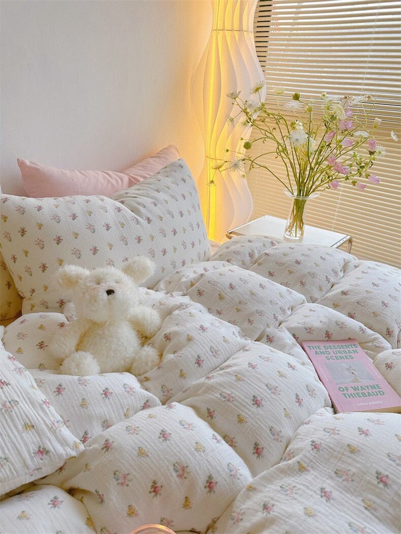 May include: A white bed with a floral patterned duvet cover and pillows. A white stuffed bunny sits on the bed. A book titled "The Deserts and Urban Scenes of Wayne Thiebaud" is on the bed.