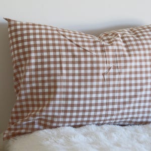 May include: A brown and white gingham pattern pillow on a white fluffy surface. The pillow is rectangular and has a classic check design. The pillow is a home decor item.
