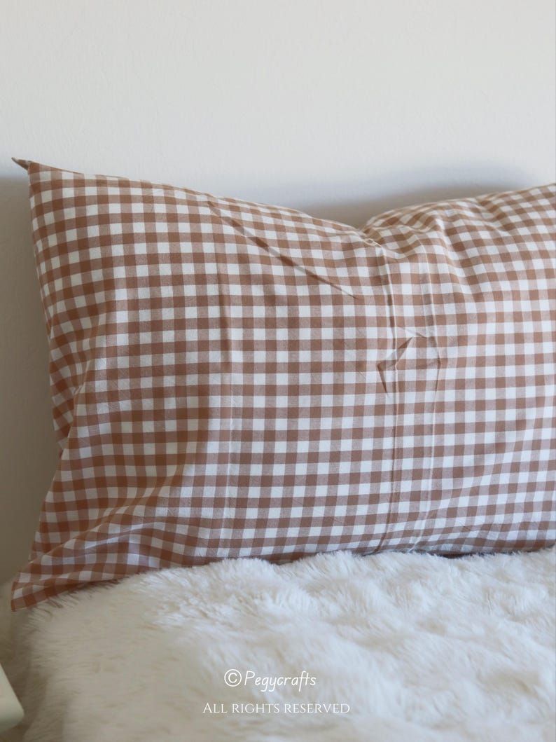 May include: A brown and white gingham pattern pillow on a white fluffy surface. The pillow is rectangular and has a classic check design. The pillow is a home decor item.