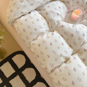 May include: A white bed with a floral patterned duvet cover and a white pillow. A lit candle sits on a white coaster on the bed. A black and white patterned rug is on the floor next to the bed.