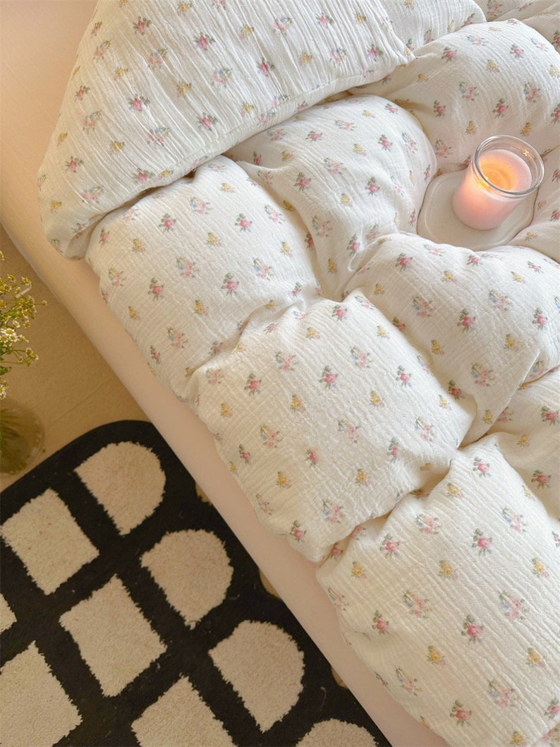 May include: A white bed with a floral patterned duvet cover and a white pillow. A lit candle sits on a white coaster on the bed. A black and white patterned rug is on the floor next to the bed.