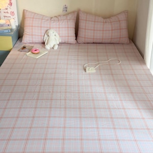 May include: A bed set featuring a light pink and white plaid pattern on the fitted sheet and two matching rectangular pillows. A white plush rabbit toy, a small book with a pink donut on top, and a white beaded handbag rest on the bed surface.