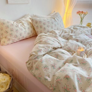 Puede incluir: Una cama rosa con un edred&oacute;n de estampado floral blanco y fundas de almohada a juego. La cama est&aacute; hecha con una s&aacute;bana bajera rosa y tiene una almohada de apoyo de estampado floral blanco encima del edred&oacute;n. Hay una vela blanca sobre un posavasos blanco en la cama.