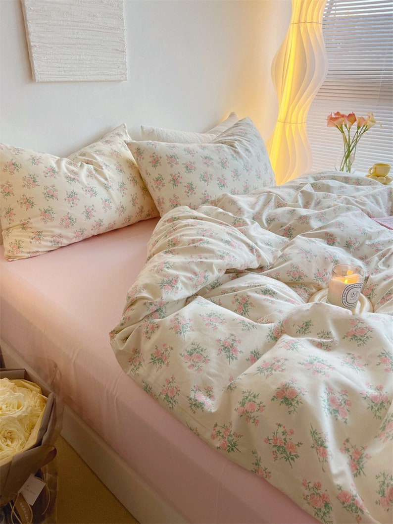 Puede incluir: Una cama rosa con un edred&oacute;n de estampado floral blanco y fundas de almohada a juego. La cama est&aacute; hecha con una s&aacute;bana bajera rosa y tiene una almohada de apoyo de estampado floral blanco encima del edred&oacute;n. Hay una vela blanca sobre un posavasos blanco en la cama.