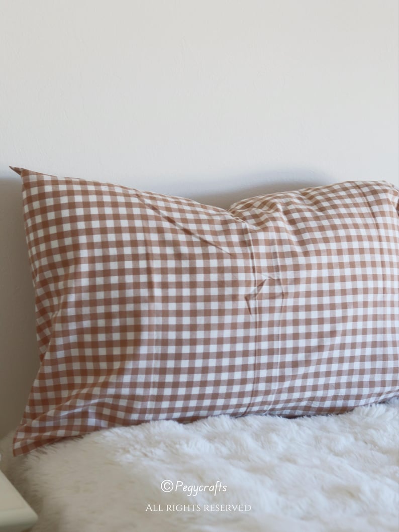 May include: A brown and white gingham patterned pillow. The pillow is rectangular and rests on a white, fluffy surface. The pillow is a warm, neutral color and would complement a variety of decor styles. The image includes the text "Pegycrafts" and "ALL RIGHTS RESERVED".