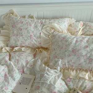 Peut inclure: Un lit avec une parure de lit à motifs floraux. La housse de couette et les oreillers présentent des roses roses et des feuilles vertes sur fond blanc. Un haut en dentelle blanche, un livre et une tasse à nœuds sont sur le lit.