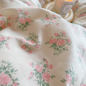 Puede incluir: Bedding blanco con un delicado estampado floral rosa. La ropa de cama es suave y lisa, con un brillo sutil. Una vela est&aacute; sentada en un posavasos blanco en el fondo.