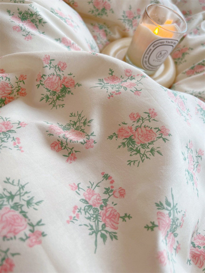 Puede incluir: Bedding blanco con un delicado estampado floral rosa. La ropa de cama es suave y lisa, con un brillo sutil. Una vela est&aacute; sentada en un posavasos blanco en el fondo.