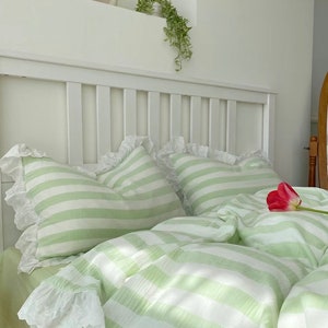 May include: A green and white striped bedspread with white lace trim. The bedspread is on a white bed frame with a white headboard. There is a single red tulip on the bedspread.