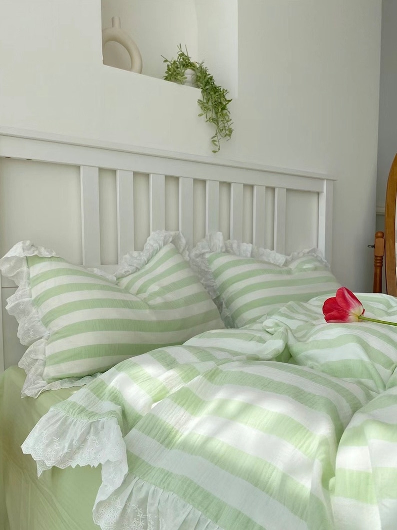 May include: A green and white striped bedspread with white lace trim. The bedspread is on a white bed frame with a white headboard. There is a single red tulip on the bedspread.
