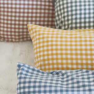 May include: A collection of gingham-patterned throw pillows in various colors, including brown, green, yellow, and blue. The pillows are arranged on a soft, white surface. The image features a close-up view of the pillows, highlighting the fabric's texture and the classic check design.