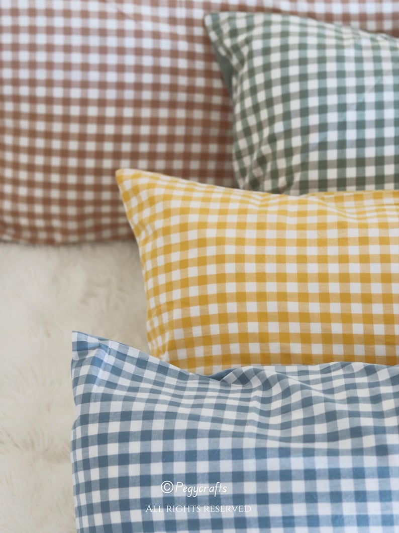 May include: A collection of gingham-patterned throw pillows in various colors, including brown, green, yellow, and blue. The pillows are arranged on a soft, white surface. The image features a close-up view of the pillows, highlighting the fabric's texture and the classic check design.