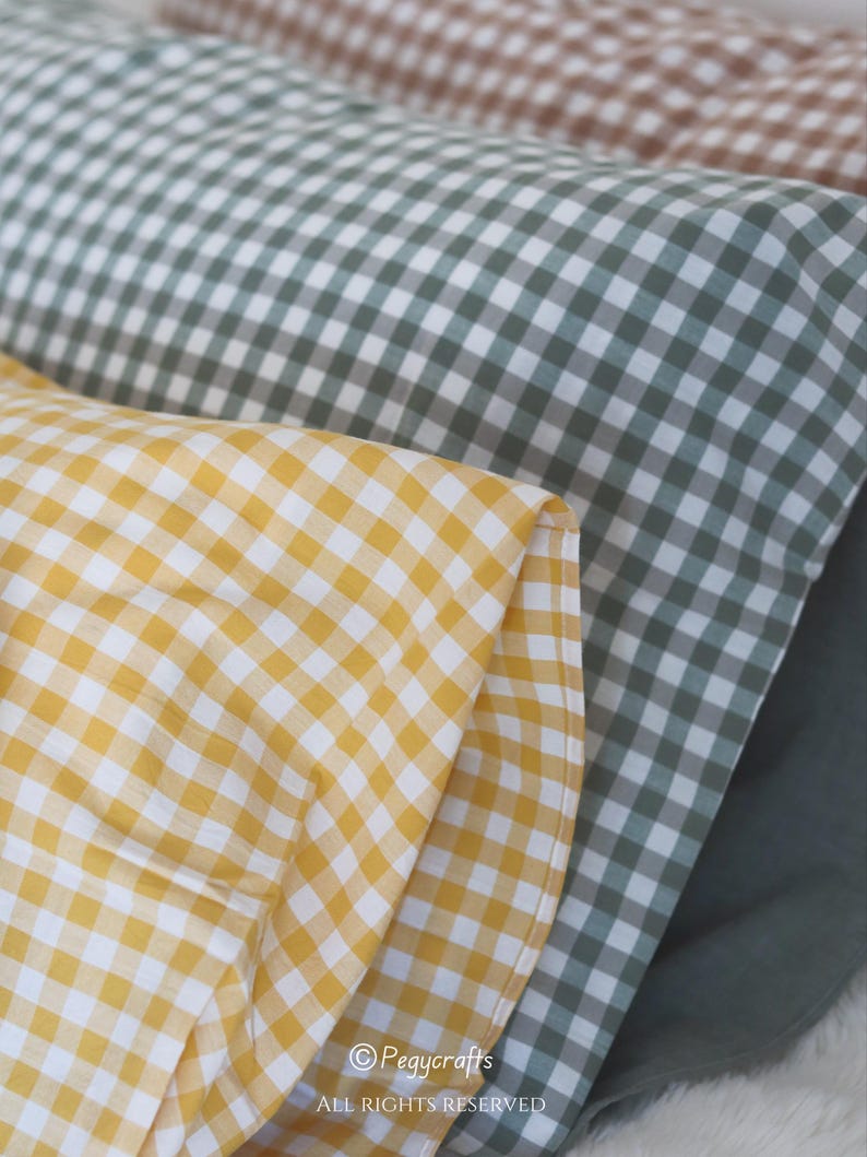 May include: Close-up of three rolls of fabric in yellow, green, and brown gingham patterns. The yellow fabric is in the foreground, followed by green and then brown. The fabric is folded and layered, showing the texture and color variations.