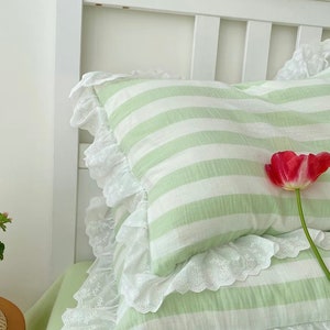 May include: Two green and white striped pillowcases with white lace trim. The pillowcases are on a white bed frame with a green and white striped pillow on top.