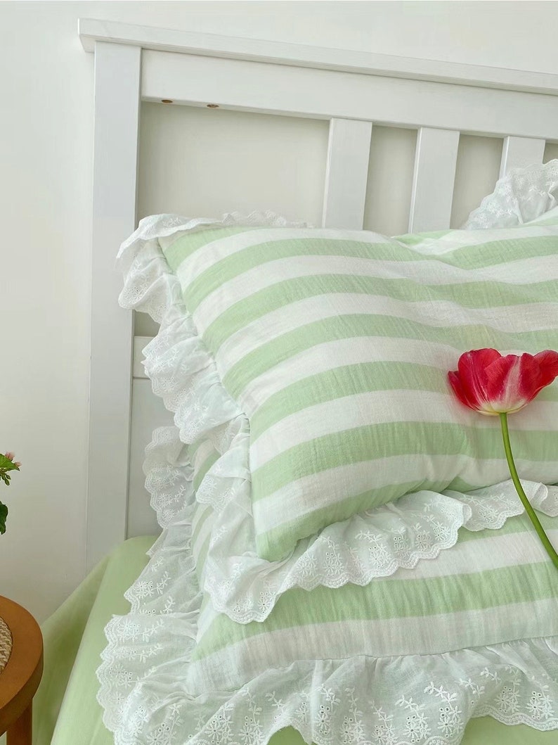 May include: Two green and white striped pillowcases with white lace trim. The pillowcases are on a white bed frame with a green and white striped pillow on top.
