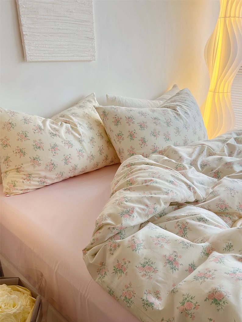 Puede incluir: Una cama rosa con un edred&oacute;n de estampado floral blanco y dos fundas de almohada a juego. La cama est&aacute; hecha con una s&aacute;bana blanca y una manta rosa.