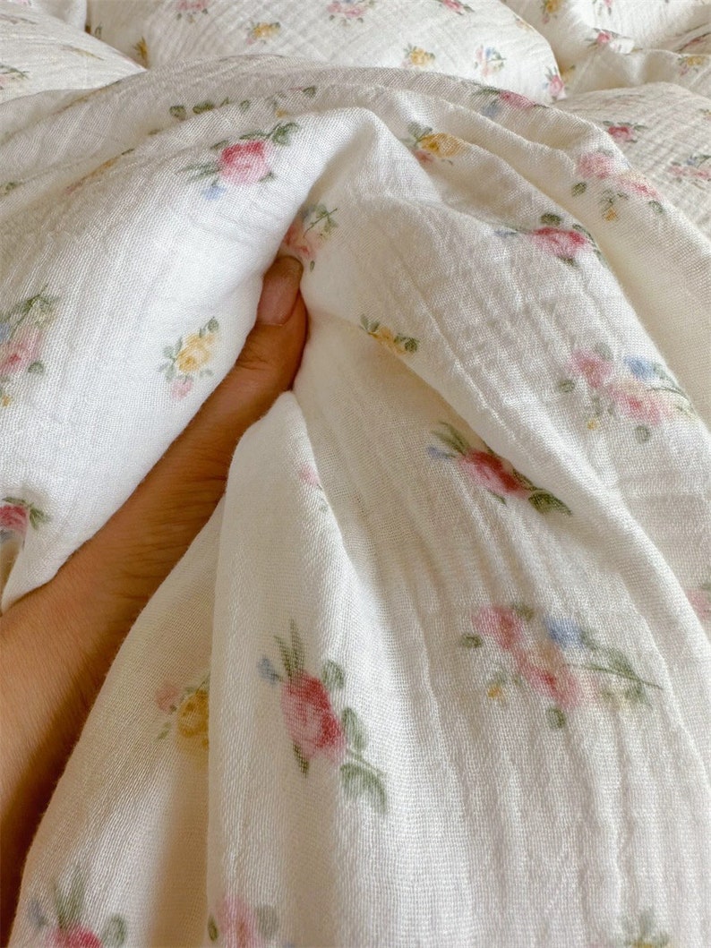 May include: White cotton fabric with a floral pattern of small pink, yellow, and blue flowers. The fabric is soft and textured, and it is perfect for making clothing, bedding, or other home decor items.