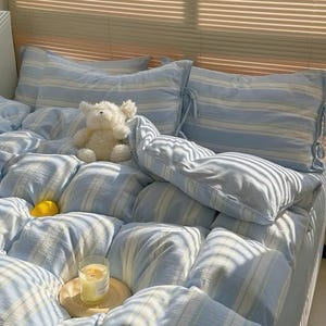 May include: A bed with light blue and white striped bedding, including pillows and a duvet. A white teddy bear and a lit candle on a wooden tray are on the bed. The word "Pegycrafts" is visible on the bottom right.