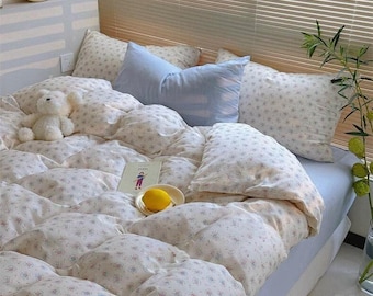 May include: A bed with a floral duvet cover and pillows. A light blue pillow is placed centrally. A small white teddy bear is on the duvet. A clock and window blinds are in the background. The scene evokes a sense of comfort.