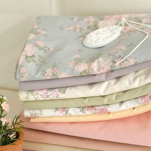 May include: A stack of folded fabric in various pastel colors, including pink, lavender, and green. The top layer features a floral pattern with pink roses on a light blue background. A white, decorative oval-shaped item rests on top.