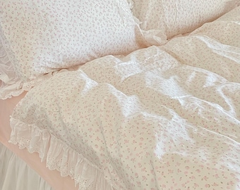 May include: A white and pink floral bedding set with white lace trim. The bedding is arranged on a bed with a white ruffled bed skirt.