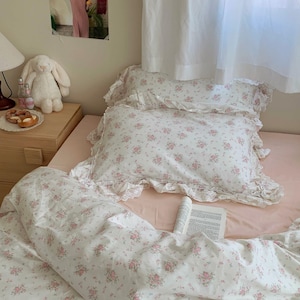 Puede incluir: Una cama blanca con un edredón y fundas de almohada con estampado floral. La cama tiene una sábana rosa y un libro encima.