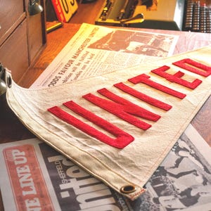 May include: A cream pennant with the word "UNITED" in red, displayed with vintage items like a clock and newspaper cuttings. The scene has a retro feel, with a "Best" sign and a wooden cabinet in the background.