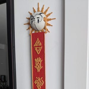Może przedstawiać: Dekoracja ścienna z motywem słońca z twarzą i czerwonym banerem. Baner ma złote symbole i złotą obwódkę. Słońce ma białą twarz z czarnymi okularami i złotym promieniem. Baner ma około 60 cm długości.