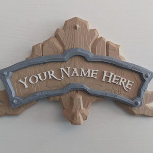 May include: A decorative wooden sign with the text "YOUR NAME HERE" in white lettering. The sign has a light brown background with a gray border and a textured wood-grain design. The sign is mounted on a light-colored surface.