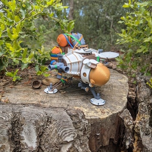 May include: A detailed model of a futuristic spacecraft, featuring a white and gold body with blue and orange accents. The model is set on a weathered tree stump, surrounded by green foliage, suggesting an outdoor setting.