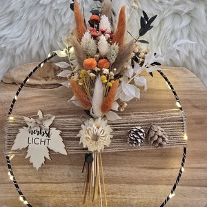 Könnte beinhalten: Ein Trockenblumenarrangement mit orangefarbenen, weißen und braunen Trockenblumen und Gräsern. Das Arrangement befindet sich in einem schwarzen Metallreifen mit einer Lichterkette. Der Reifen liegt auf einer Holzoberfläche. Der Text "herbst LICHT" befindet sich auf einem Holzblatt.