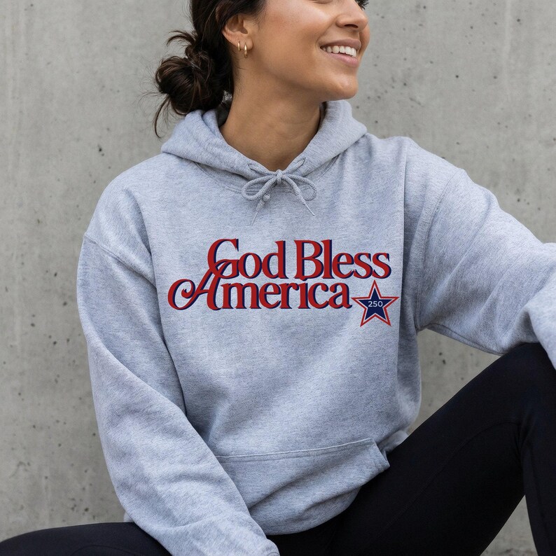 May include: A light gray hoodie with the text "God Bless America" in red and blue script, accompanied by a blue star graphic. The hoodie features a drawstring and a kangaroo pocket.