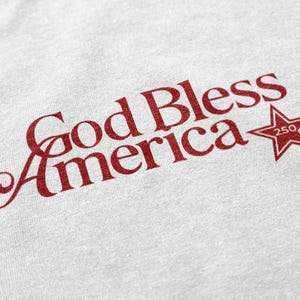 May include: A white t-shirt with the phrase "God Bless America" in red script. A red star with the number "250" is next to the text. The shirt has a slightly textured appearance.