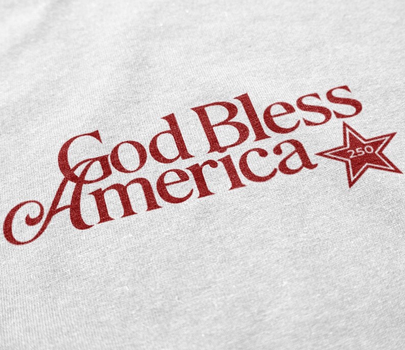 May include: A white t-shirt with the phrase "God Bless America" in red script. A red star with the number "250" is next to the text. The shirt has a slightly textured appearance.