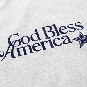 May include: Close-up of a light gray fabric with the navy blue text "God Bless America" in a script font. A navy blue star with the number "250" inside is to the right of the text. The fabric has a subtle texture.
