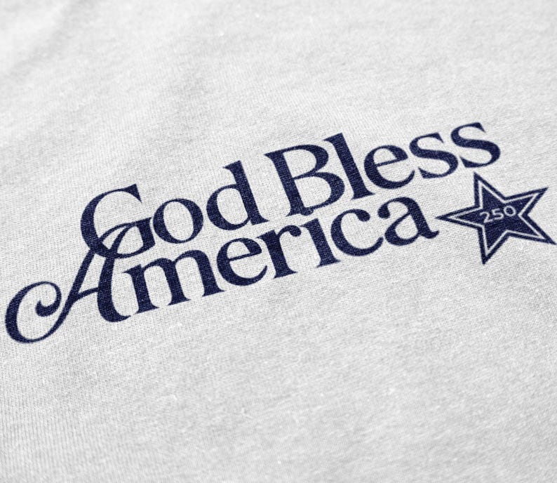 May include: Close-up of a light gray fabric with the navy blue text "God Bless America" in a script font. A navy blue star with the number "250" inside is to the right of the text. The fabric has a subtle texture.