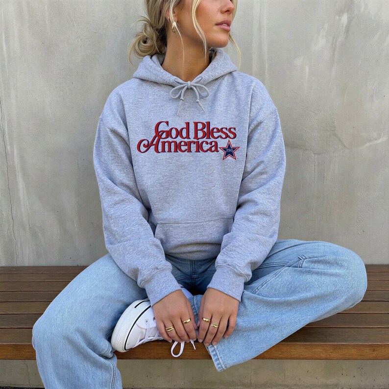 May include: A gray hoodie with the text "God Bless America" in red and blue, featuring a star graphic. The hoodie has a drawstring and is paired with light blue jeans and white sneakers.