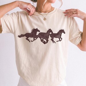 Puede incluir: Una camiseta de manga corta color crema con un estampado gráfico marrón oscuro de tres caballos corriendo en línea por el centro. La camiseta tiene cuello redondo y se lleva con un collar de cadenas doradas superpuestas con colgante.