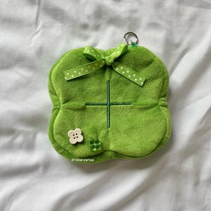 May include: A green plush four-leaf clover keychain with a white button and green glitter accents. The clover has a white ribbon with green polka dots tied around it.