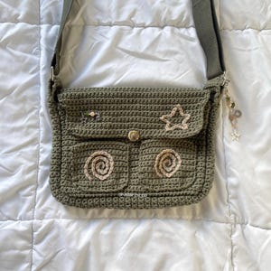 May include: Olive green crochet shoulder bag with a flap closure and two front pockets. The bag features a star and spiral design, with a matching strap and decorative charms. The bag is handmade.