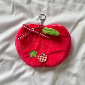 May include: A bright pink plush apple keychain with a red and white gingham bow, a green leaf, and a star and apple button detail.  The keychain has a silver metal ring.