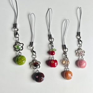 May include: Five colorful phone charms with different designs. Each charm features a small, round, 3D object, such as a green fruit, a red apple, a brown chocolate, a pink donut, and an orange pastry. Each charm is attached to a silver chain and a thin, gray string.